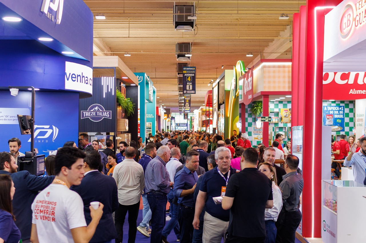 Corredor lotado da ExpoApras com estandes de marcas de alimentos, sinalização de avenidas e ruas do pavilhão