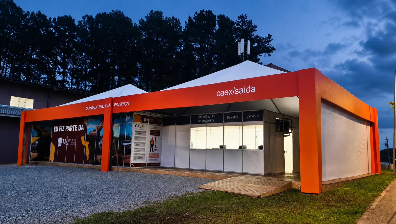 CAEX e credenciamento da Arena M&T 2026 ao entardecer, com cobertura branca e estrutura laranja, painéis de comunicação visual e balcões de atendimento iluminados