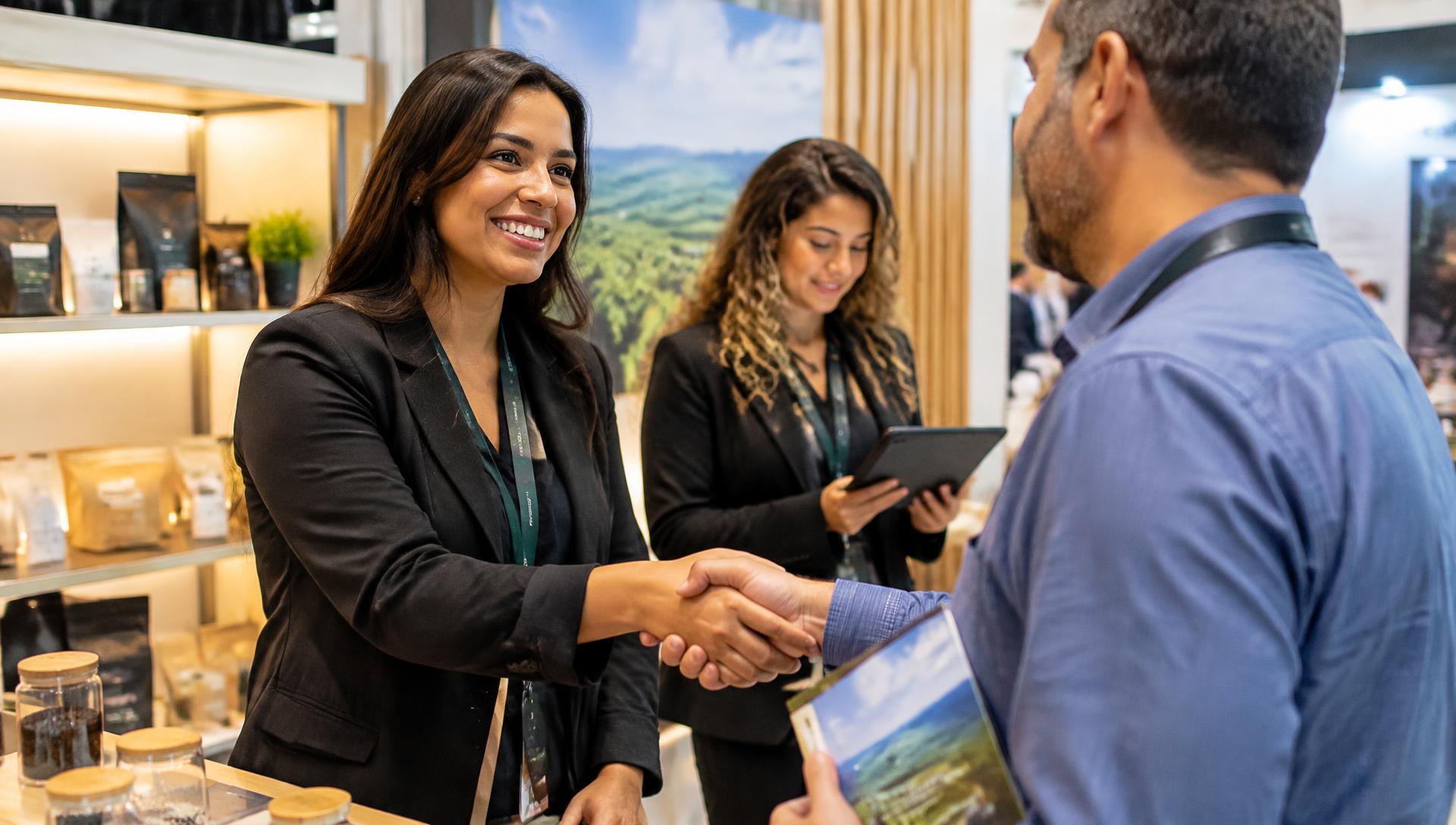Vendedora de stand cumprimentando visitante com aperto de mãos enquanto colega registra o contato