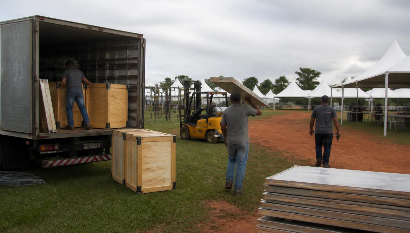 Descarregamento de caixas de madeira e painéis de stand em caminhão no parque de exposições, com empilhadeira e tendas ao fundo