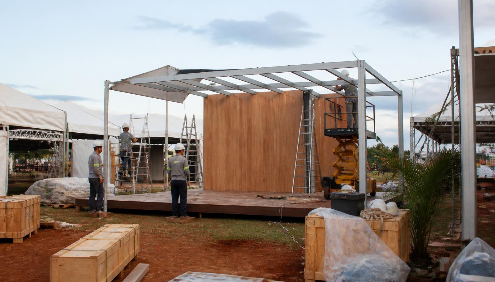 Equipe de montagem uniformizada instalando estrutura metálica e painéis de madeira de um stand em terreno aberto do parque de exposições