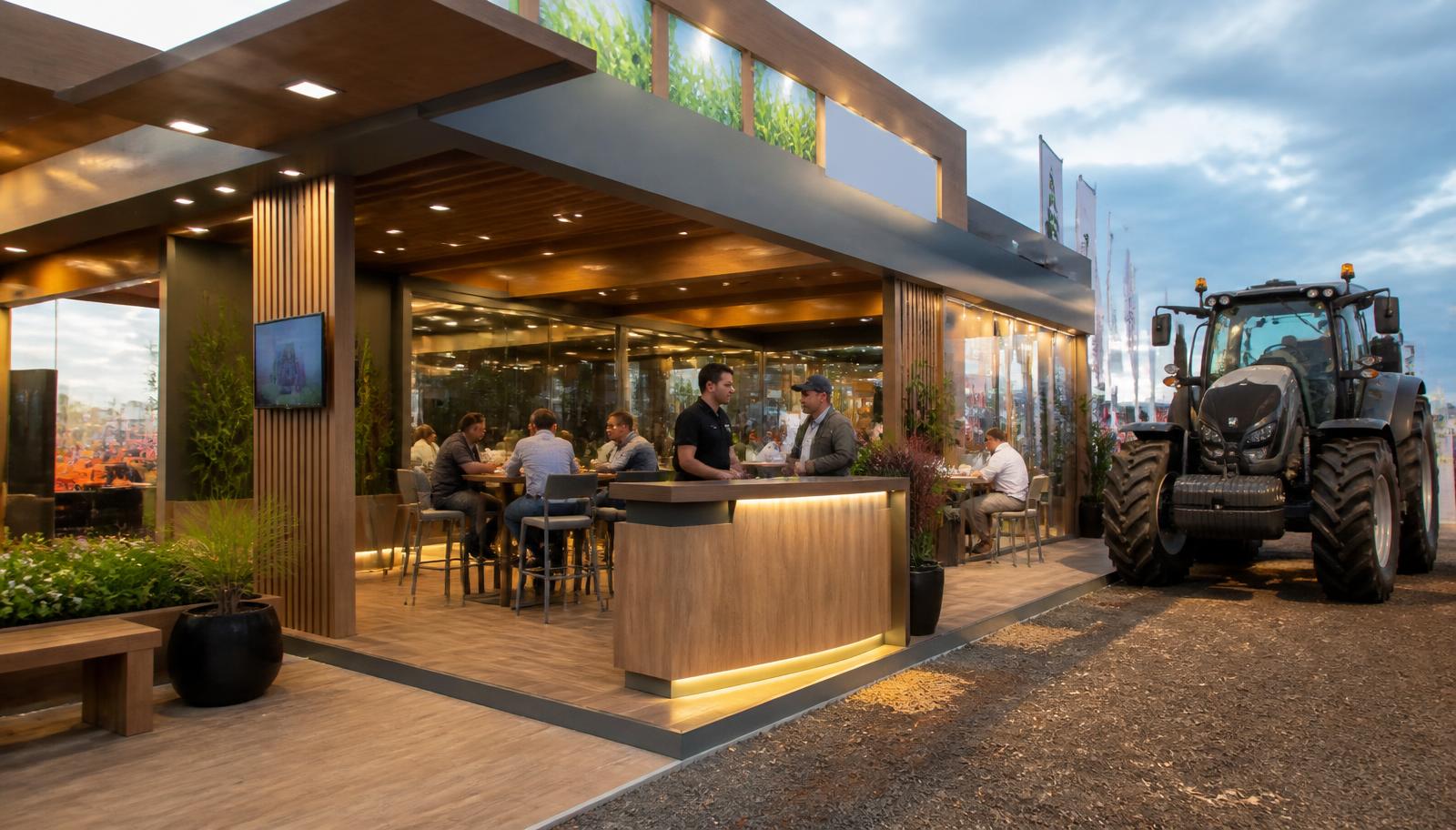 Stand construído em madeira e metal em feira agrícola a céu aberto, com balcão iluminado, sala de reunião envidraçada e trator ao lado