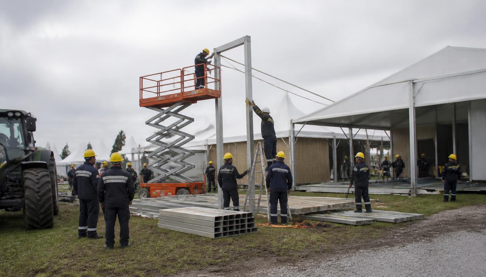 Equipe uniformizada montando estrutura modular de estande sob tenda branca em parque de exposições antes da abertura da feira