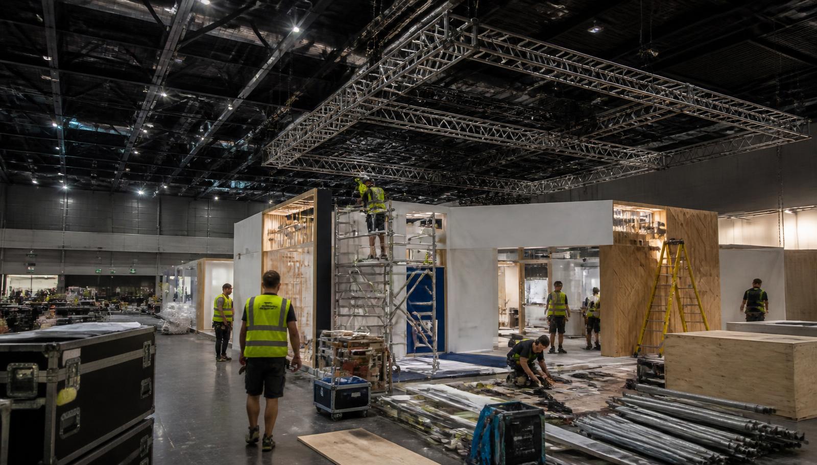 Equipe de montagem trabalhando em estruturas de stands e treliças dentro de pavilhão de exposições durante a madrugada